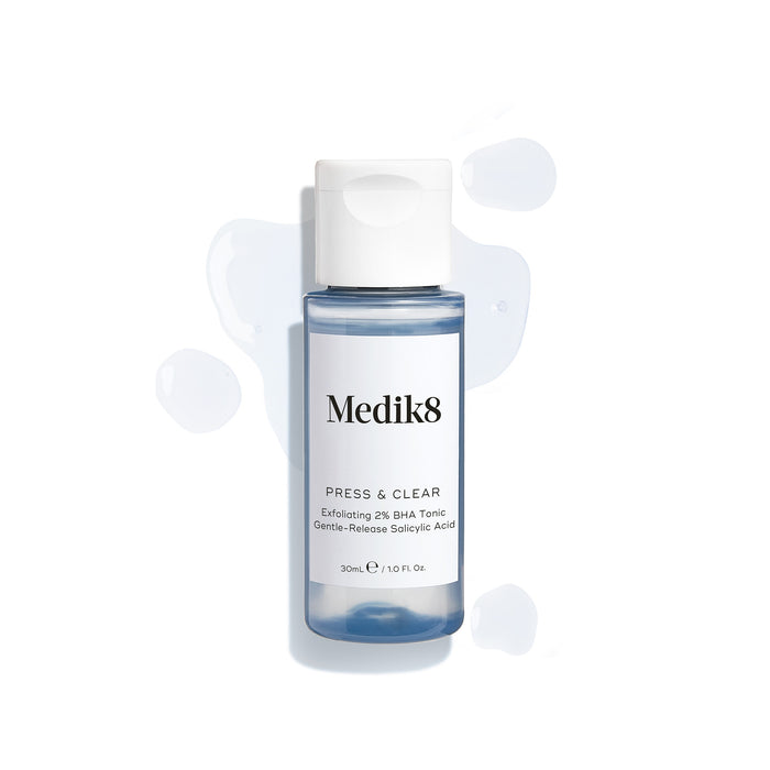 A travel-size bottle (30ml) of Medik8 Press & Clear tonic with a white flip-top cap, surrounded by clear blue droplets on a white surface. Text identifies it as an "Exfoliating 2% BHA Tonic."
